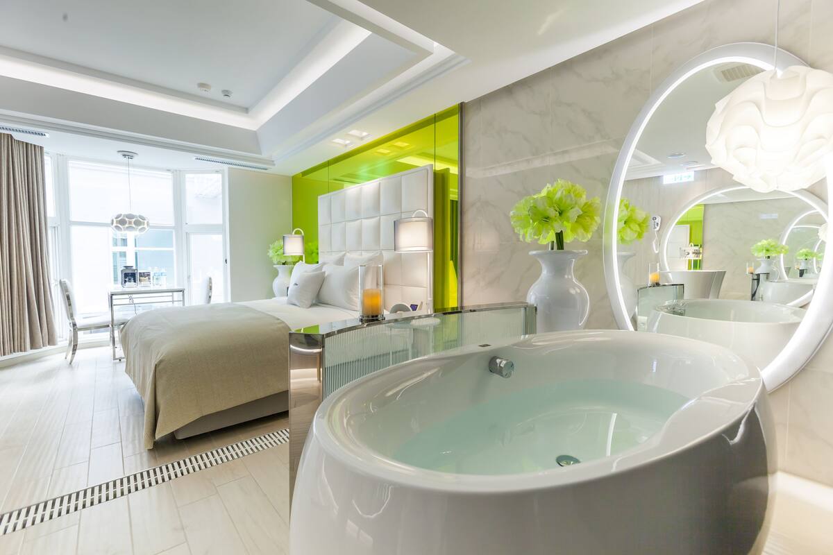 A modern hotel room with a large bed, a glass-enclosed seating area, a large mirror, and a bathtub.