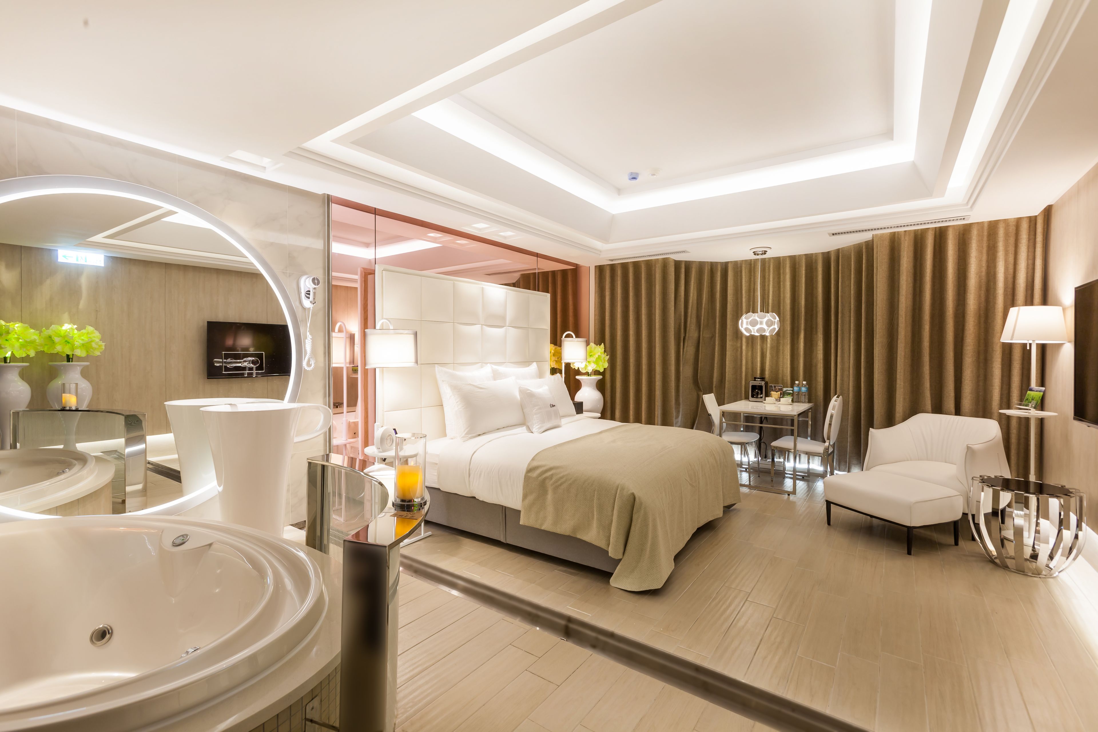 A modern hotel room with a large bed, a sitting area with a table and chairs, a separate bathroom with a large mirror and sink, and a television.
