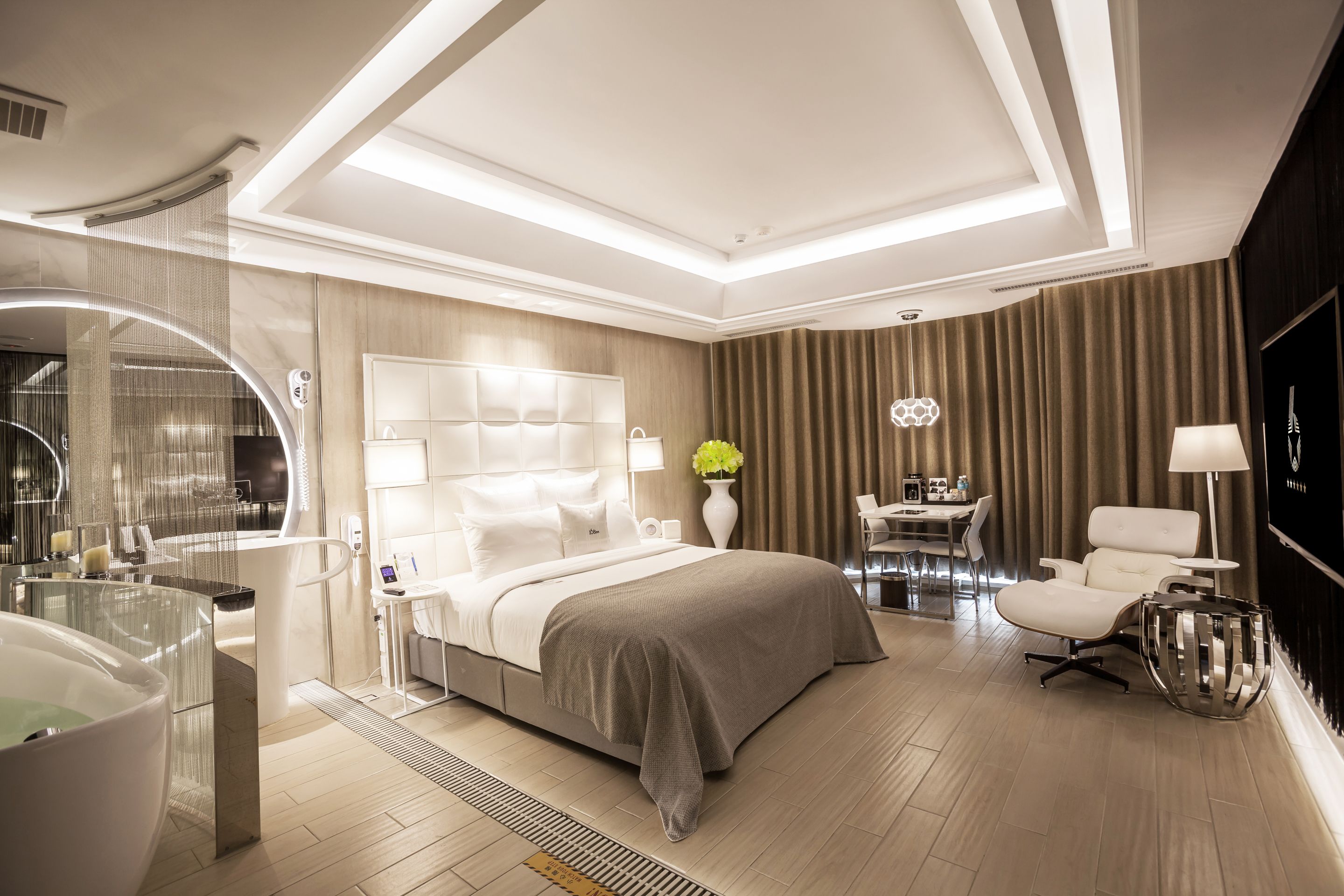 A modern hotel room with a large bed, a sitting area with a table and chairs, a large mirror, and a TV.