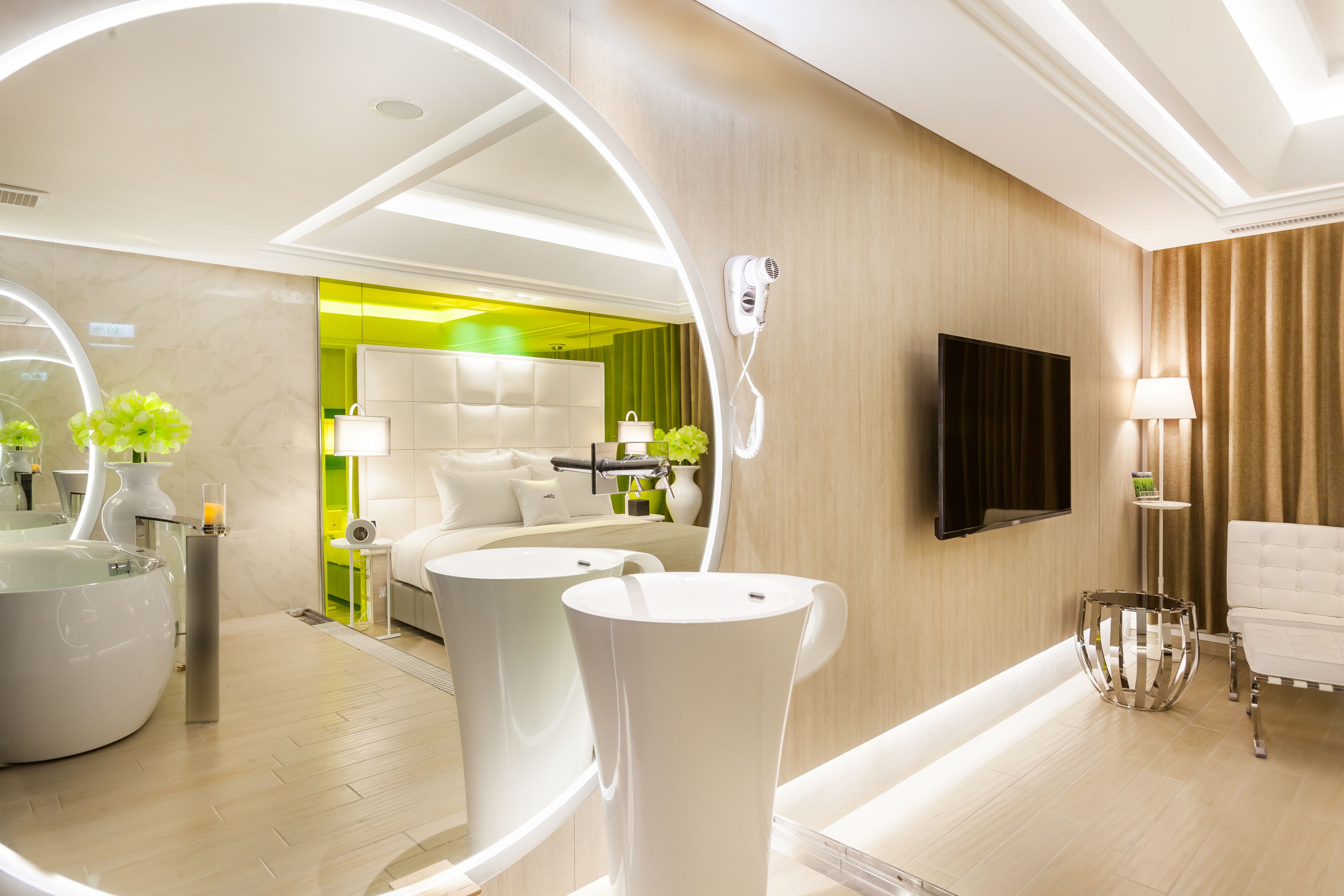 A modern hotel room with a large mirror, a round bathtub, a sofa, and a television.