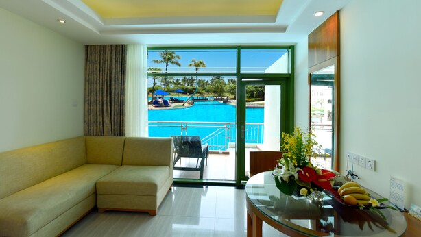 A hotel room with a sofa, a glass table with fruit and flowers, and a view of a pool and palm trees.