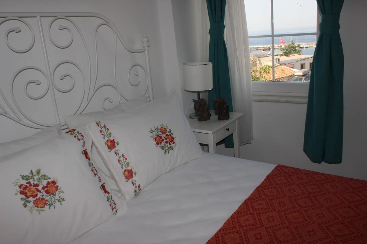 A bed with floral pillows, a red blanket, and a view of the sea through a window with curtains.