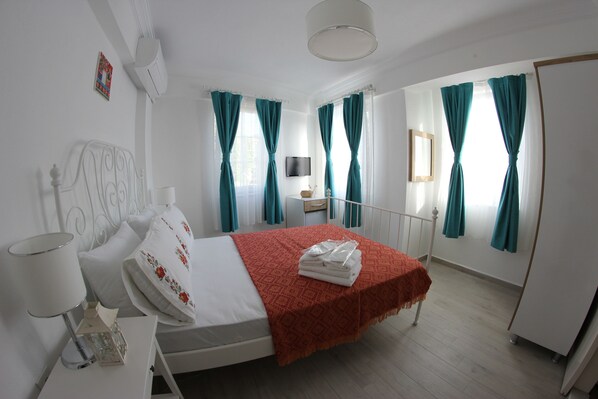 A bedroom with a bed, a bedside table with a lamp, a TV on the wall, and teal curtains. - Yeldeğirmeni Bozcaada (Bozcaada)