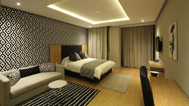 A modern hotel room with a geometric patterned wall, a bed with a black headboard, a sofa, a small round table, and a wooden bench. - My Loft Anfa (Casablanca)