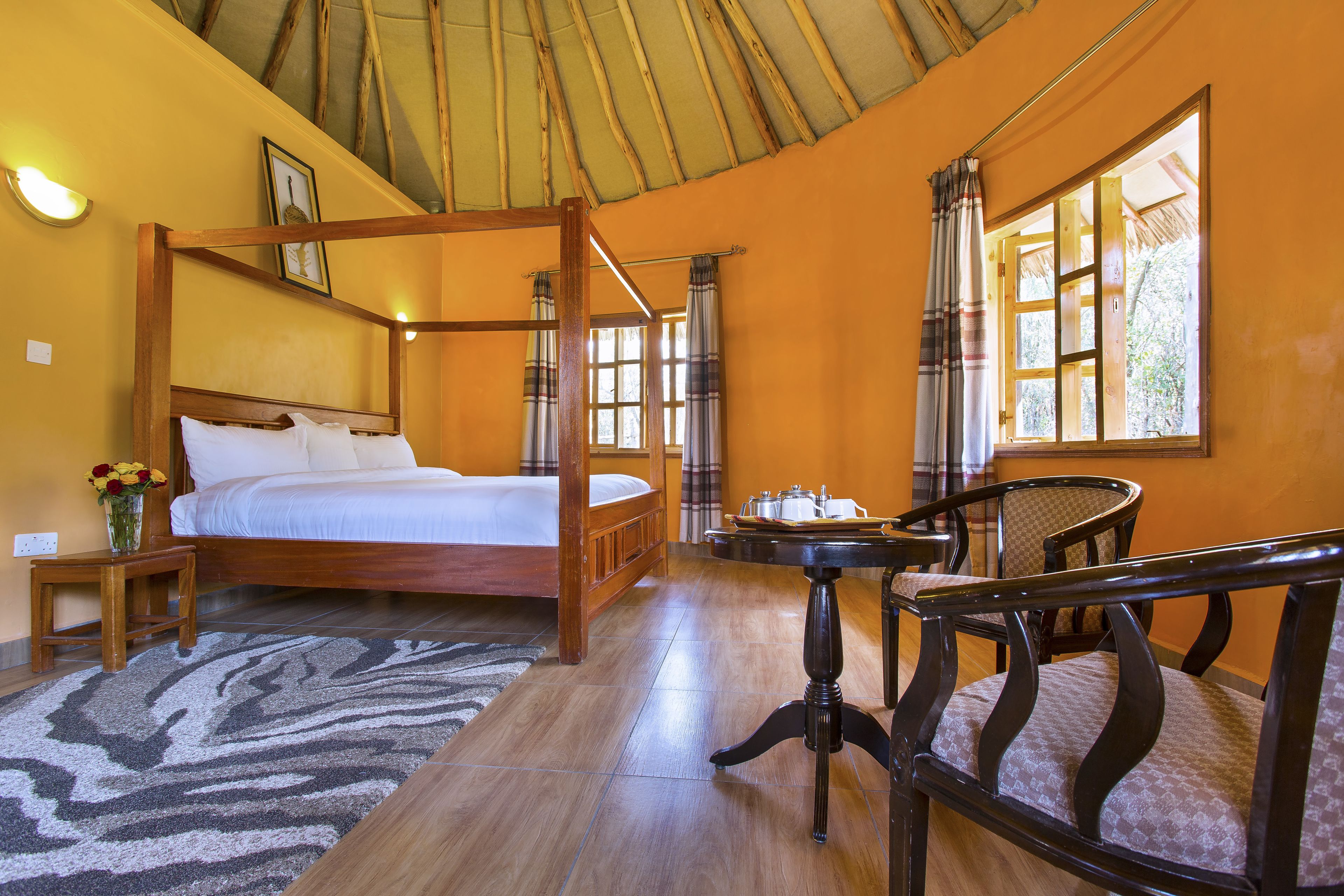 A four-poster bed with a wooden frame, a small wooden table with a tray, a chair, and a window with curtains.