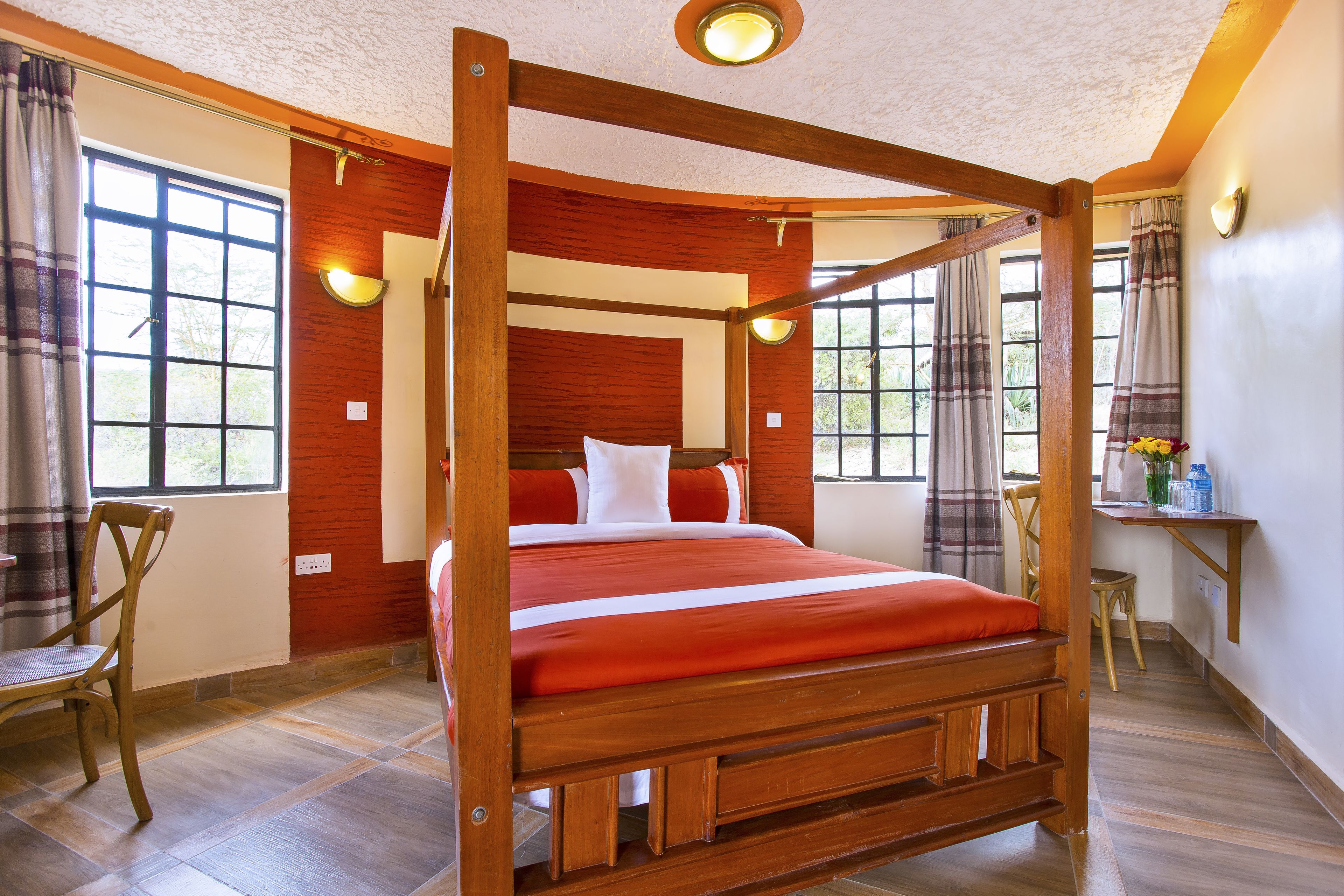 A four-poster bed with a red bedspread, wooden headboard, and white pillows. There is a wooden chair and a small table with a vase of flowers.