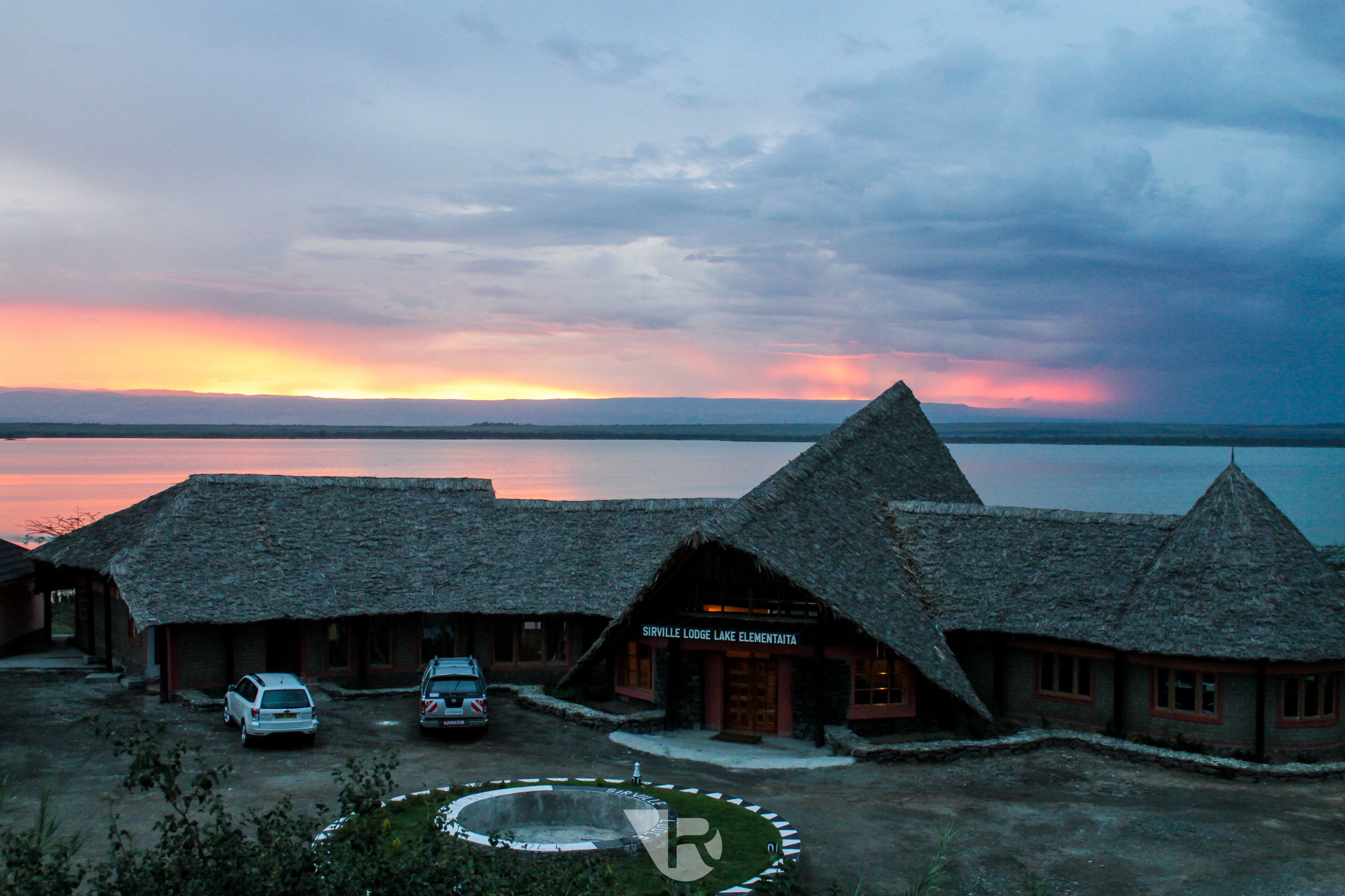 A lodge with thatched-roof buildings, a swimming pool, and a view of a large body of water at sunset.
