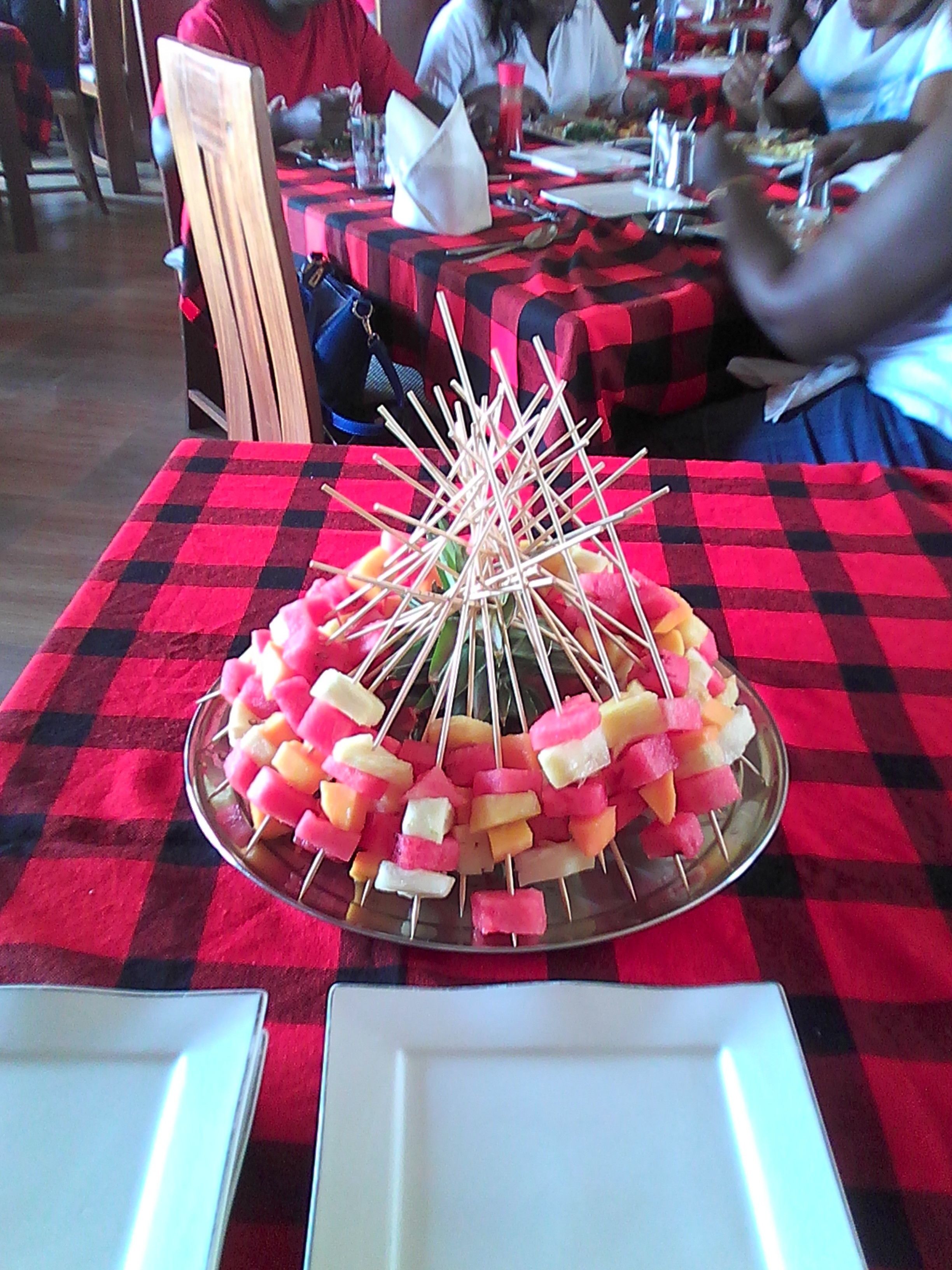 A dining table with a red and black checkered tablecloth, a centerpiece of skewered fruit, and a plate in the foreground.