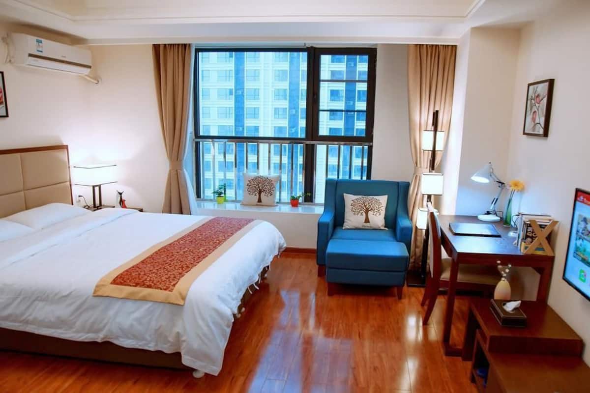 A hotel room with a large bed, a blue armchair, a desk with a computer, and a window with a city view.