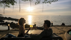 A couple toasting with champagne glasses at a wooden table with a view of the sunset over the sea. - Villa Riva (Koh Samui)