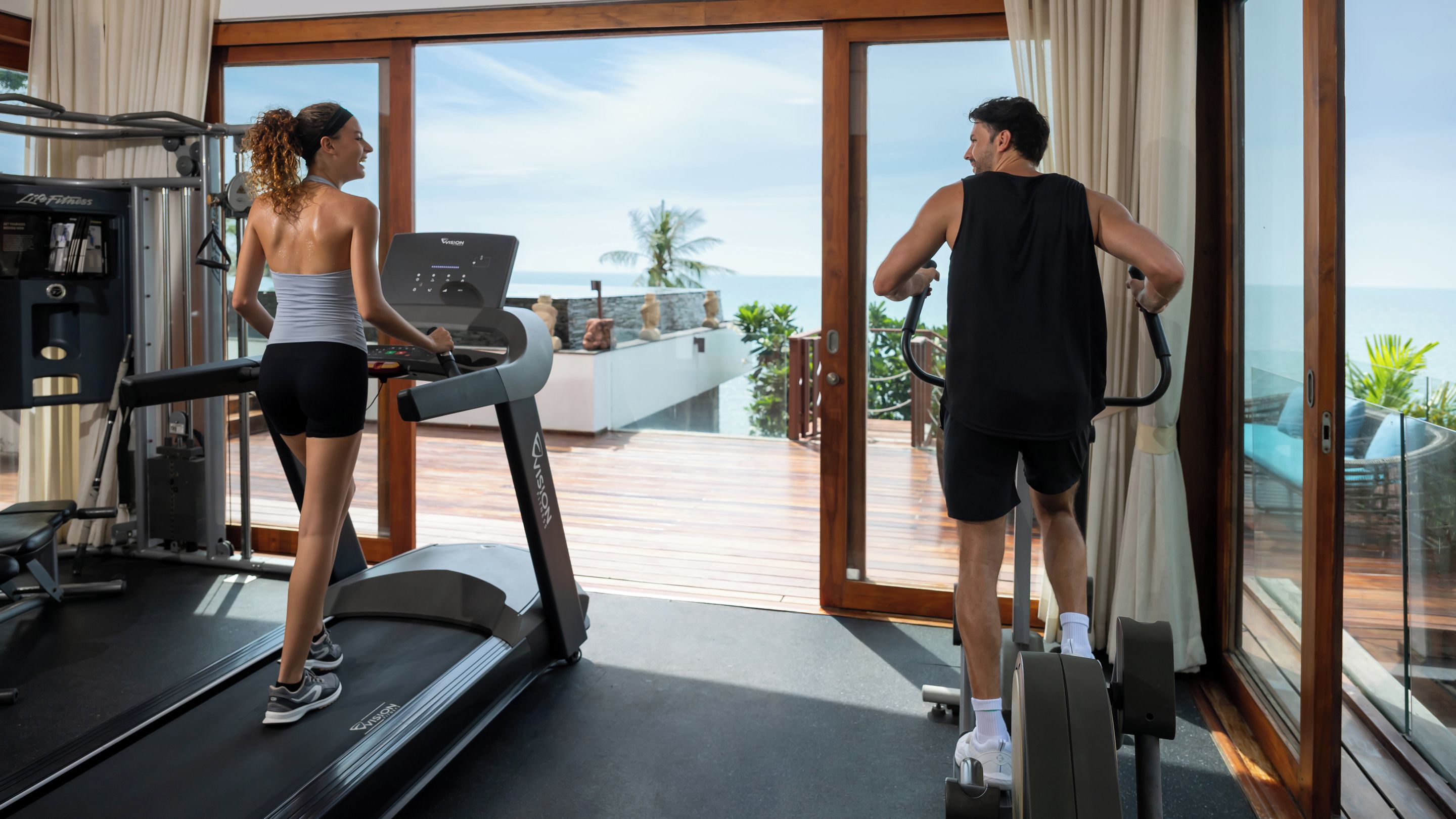 A fitness area with treadmills and exercise equipment, large windows with a view of the ocean, and a wooden deck.