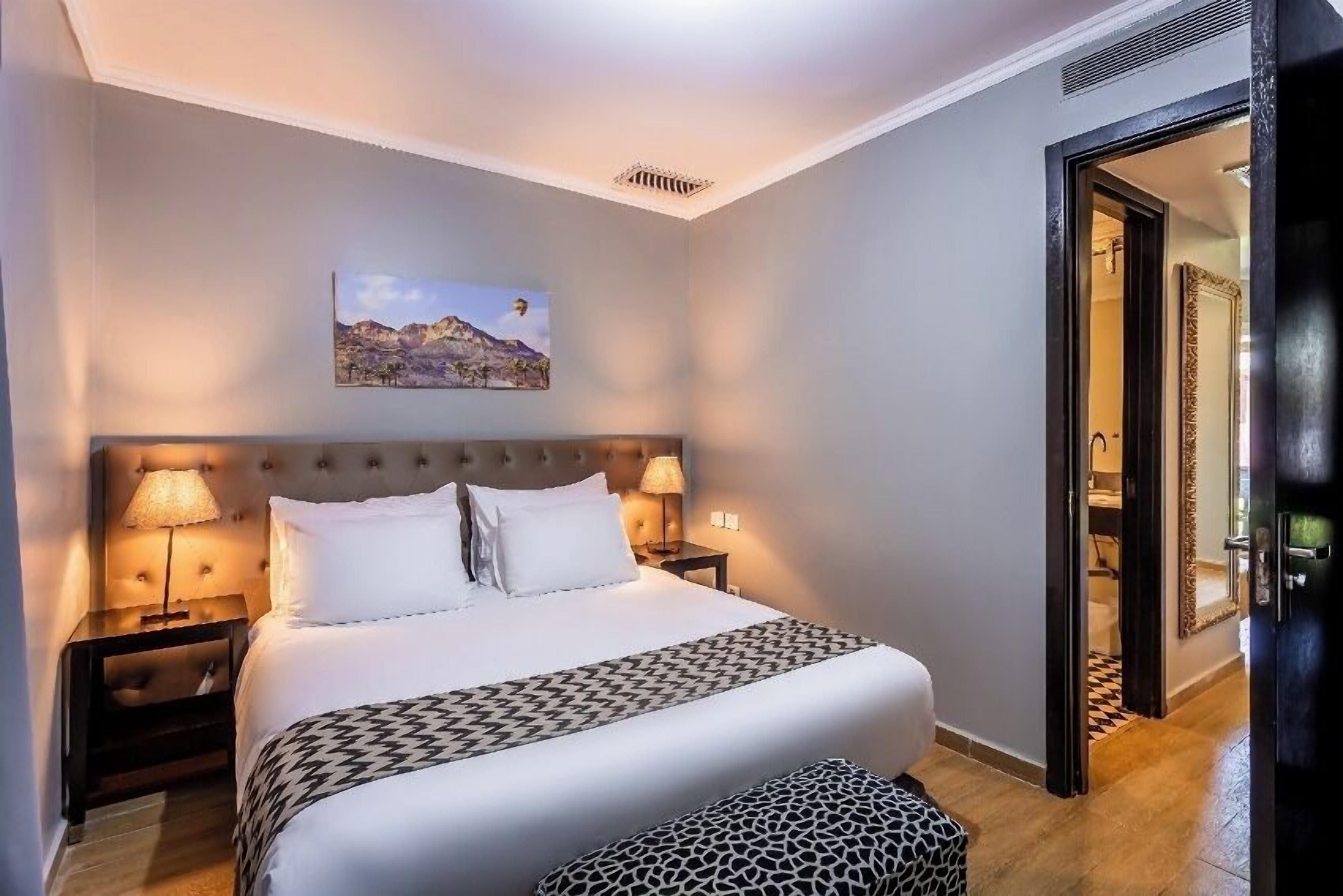 Une chambre d’hôtel avec un grand lit, deux lampes de chevet, une tête de lit, un tableau au mur et une porte donnant sur une autre pièce.
