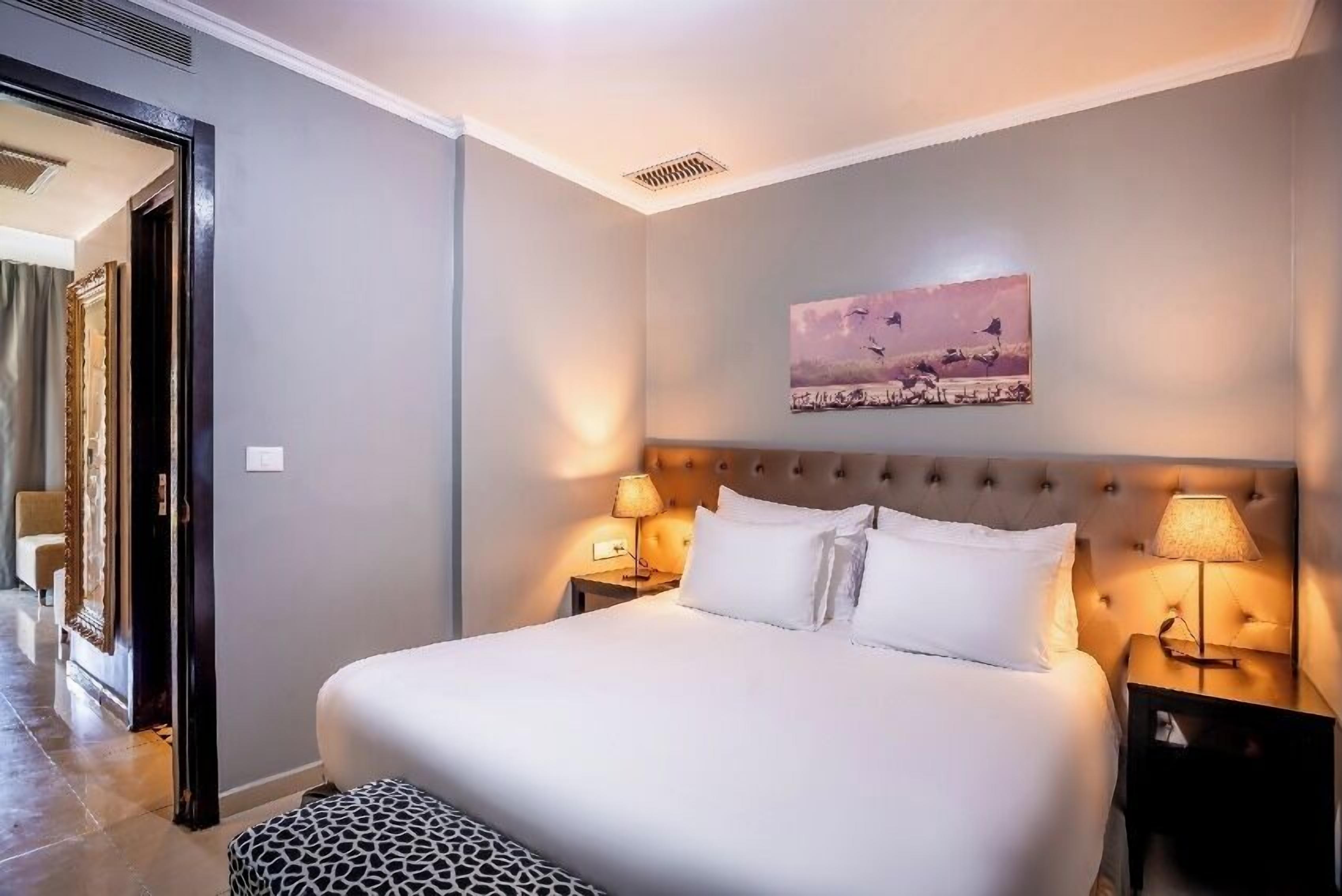 Een netjes opgemaakt bed met witte lakens, twee bedlampjes en een wanddecoratie van vogels.
