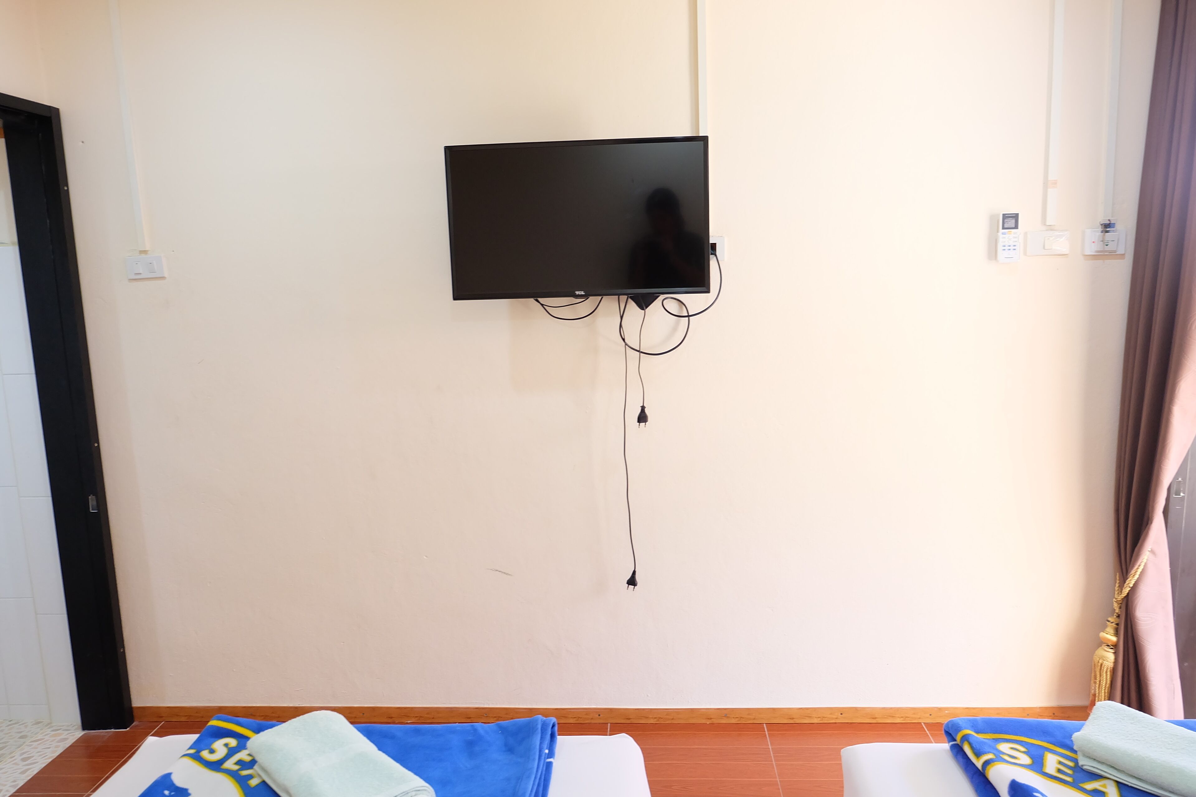Una habitación con una televisión de pantalla plana montada en la pared, una cama con una colcha azul y blanca, y piso de madera.