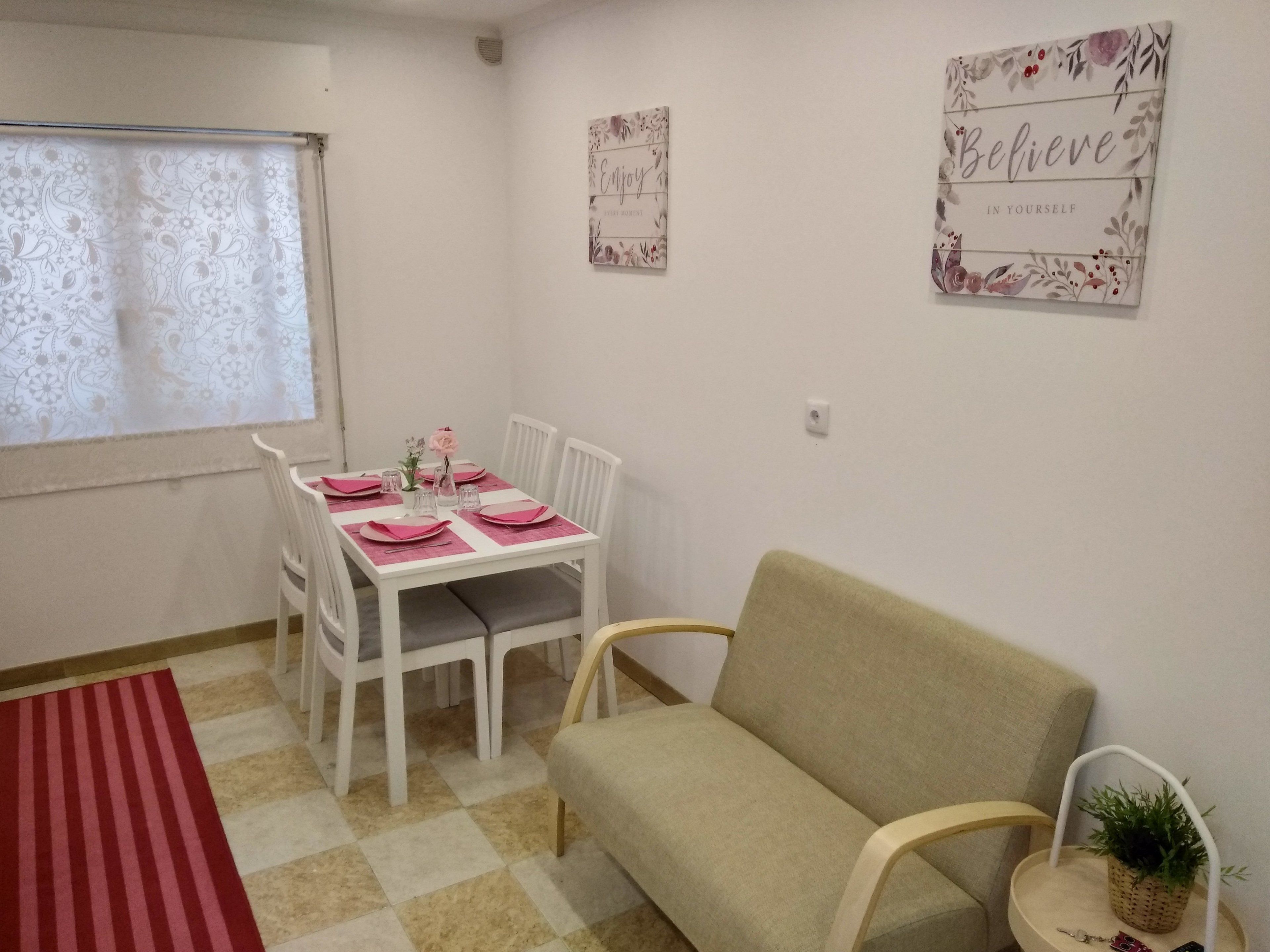 A dining area with a table set for four, white chairs, a red and beige checkered floor, and two wall art pieces.