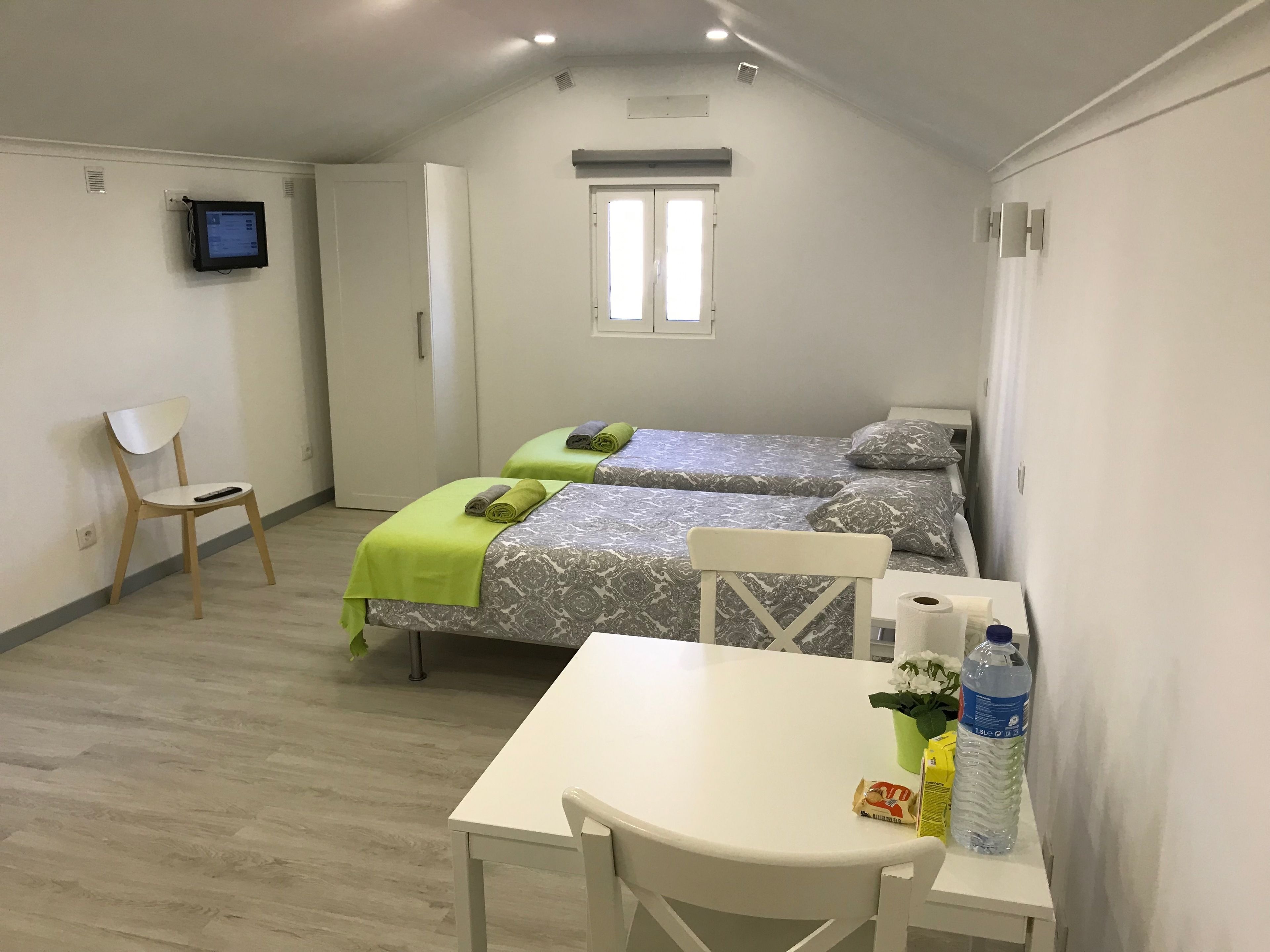 A small, well-lit room with two beds, a single chair, a table, and a TV mounted on the wall.