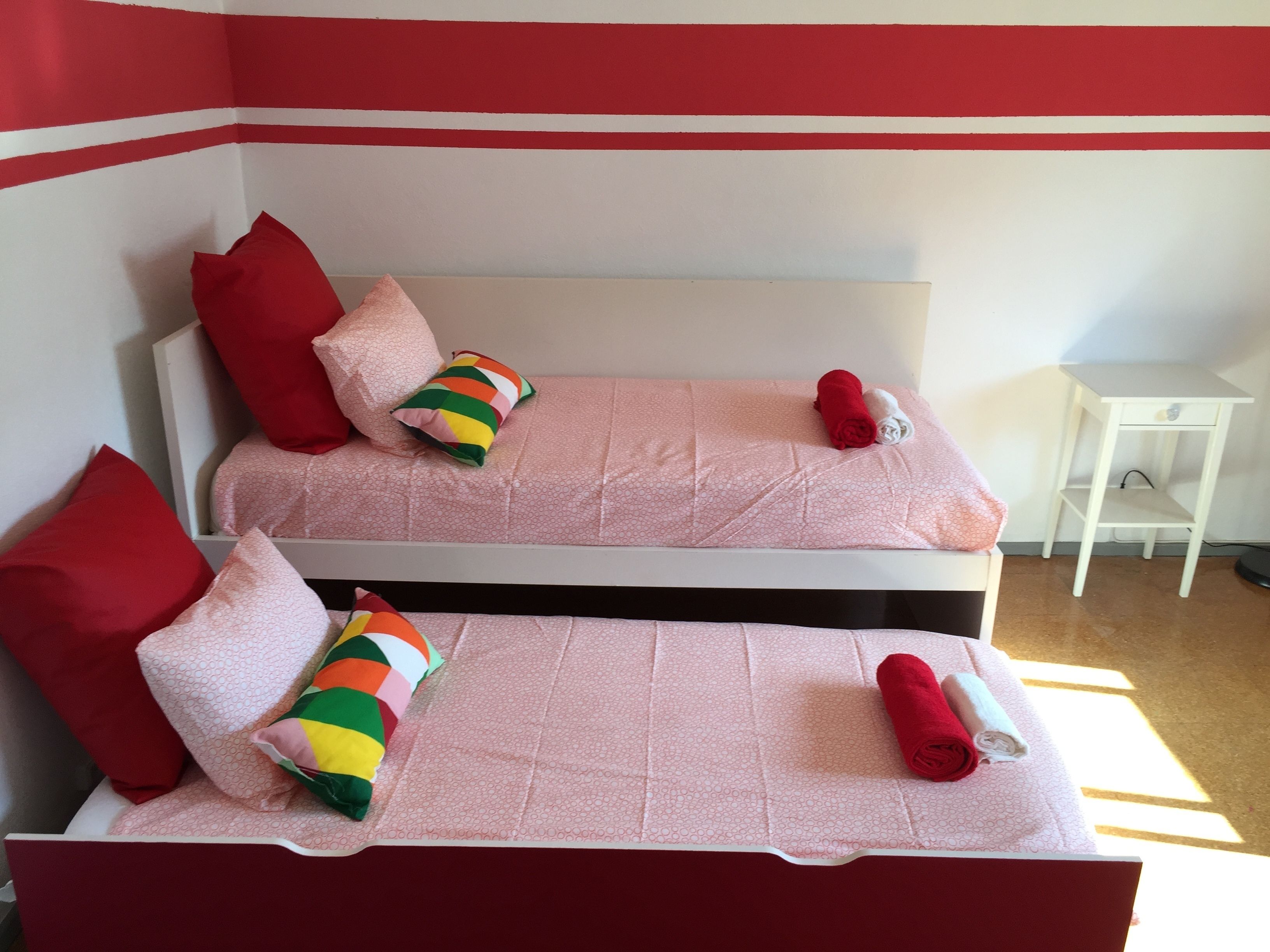 Two single beds with pink bedding and red pillows, a small white table, and a red and white striped pillow.
