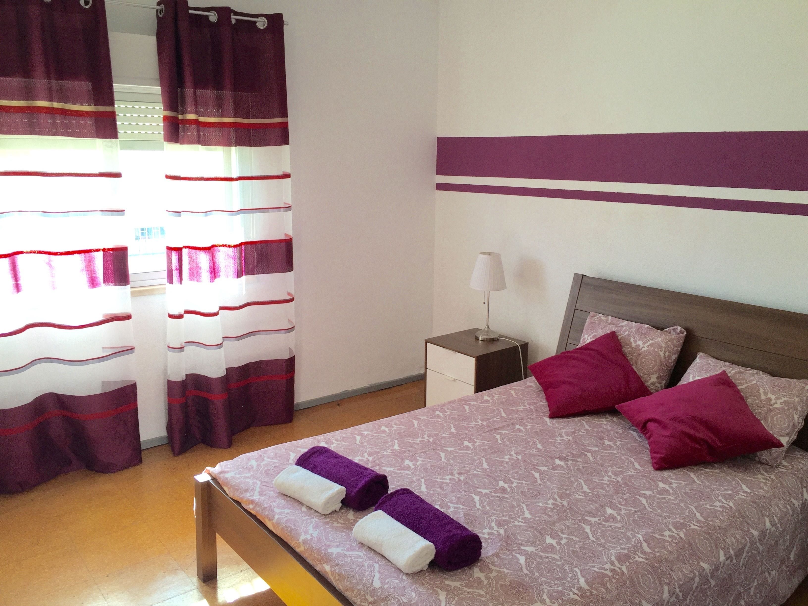 A hotel room with a bed, pillows, a nightstand, a lamp, and curtains.