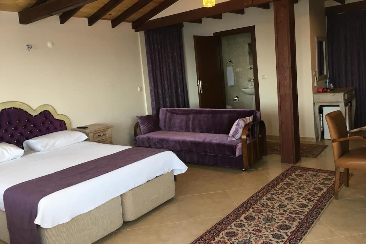 A bedroom with a bed, a purple sofa, a chair, a small table with a lamp, and a bathroom visible through an open door.