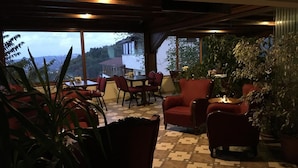 A covered outdoor seating area with red chairs and tables, surrounded by potted plants, and overlooking a building with a view of the landscape. - Dilek Konağı (Sapanca)