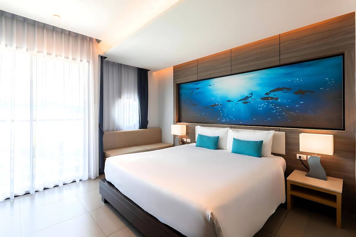 A modern hotel room with a large bed, a wall-mounted TV, and a view of the ocean through the window.