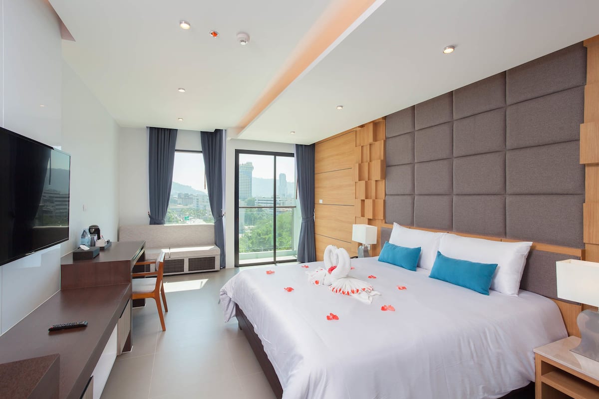 A modern hotel room with a large bed, a desk, a TV, and a balcony with a city view.