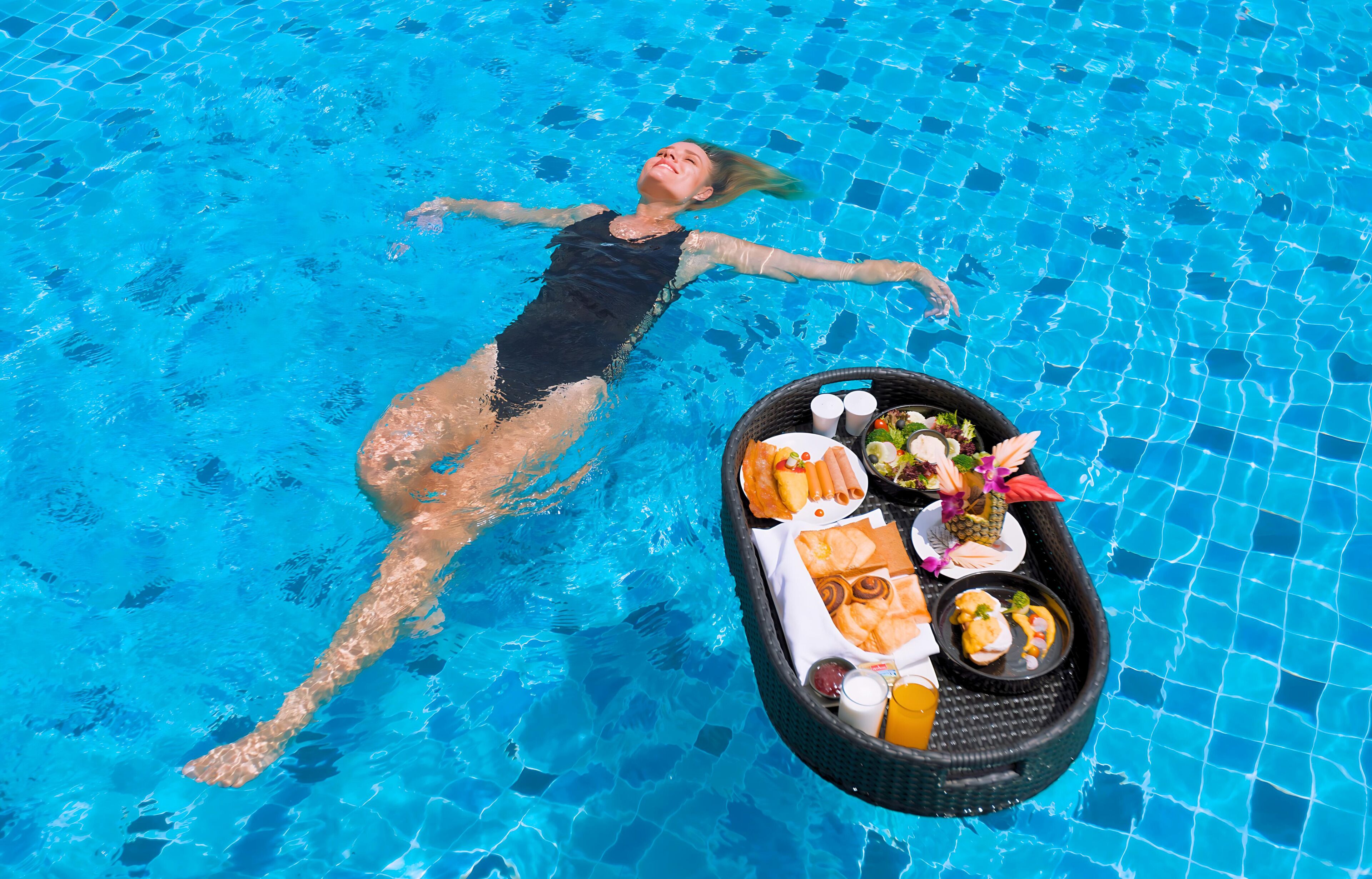 Eine Person treibt in einem Pool, auf einem schwimmenden Tablett befinden sich Speisen und Getränke.