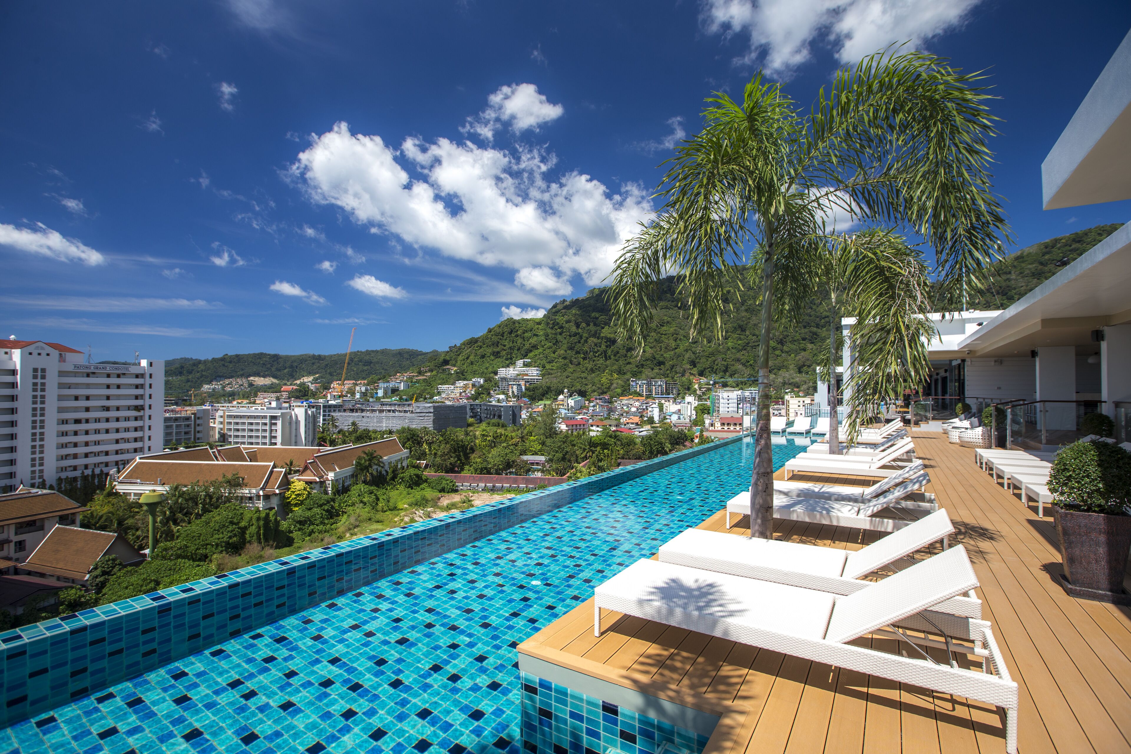 A rooftop pool with lounge chairs and palm trees, overlooking a cityscape with buildings and hills.