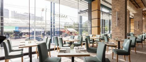 Een modern restaurant met grote ramen, groene stoelen en houten tafels.