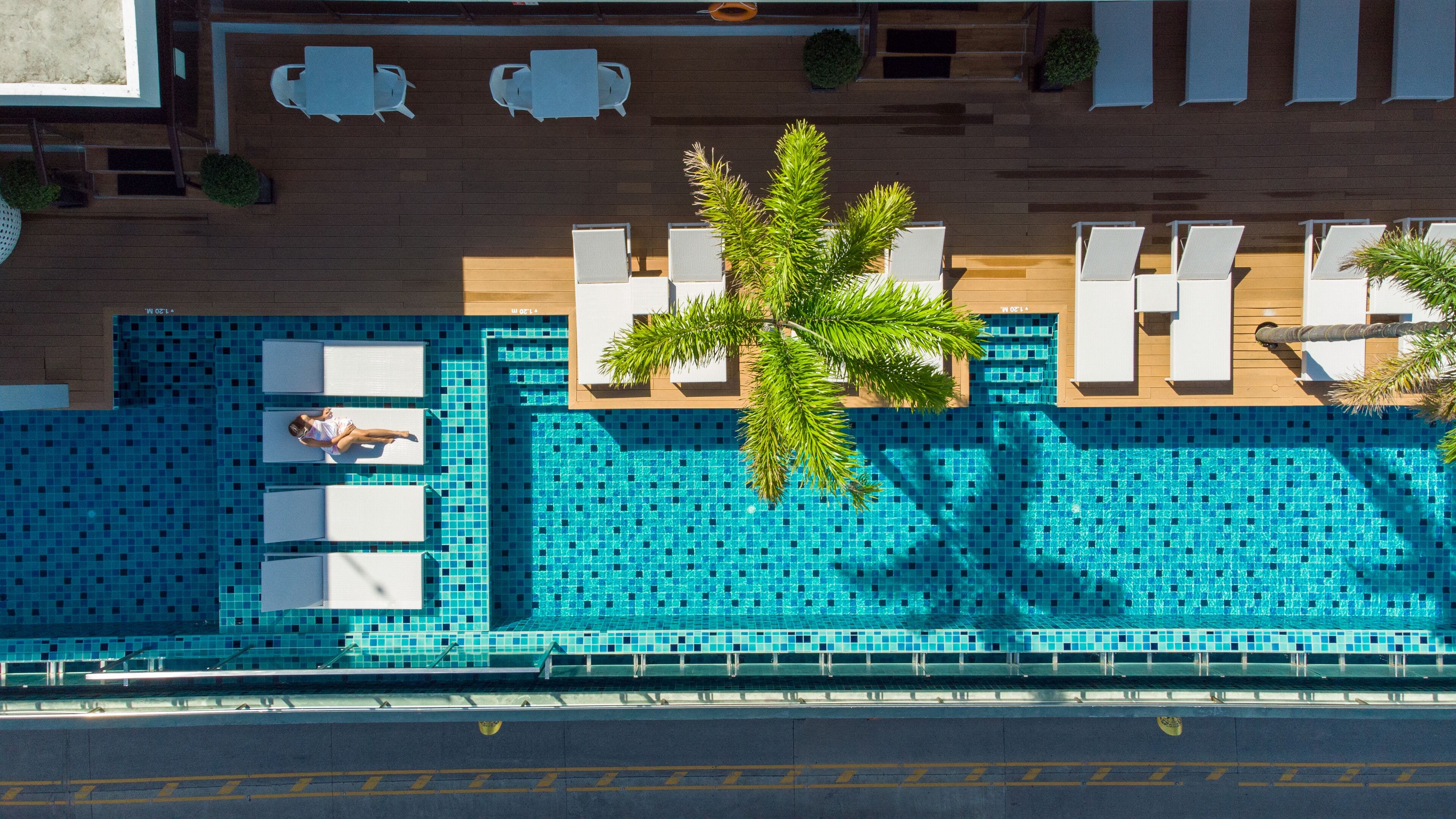 An aerial view of a hotel pool area with a tiled pool, lounge chairs, and a palm tree.