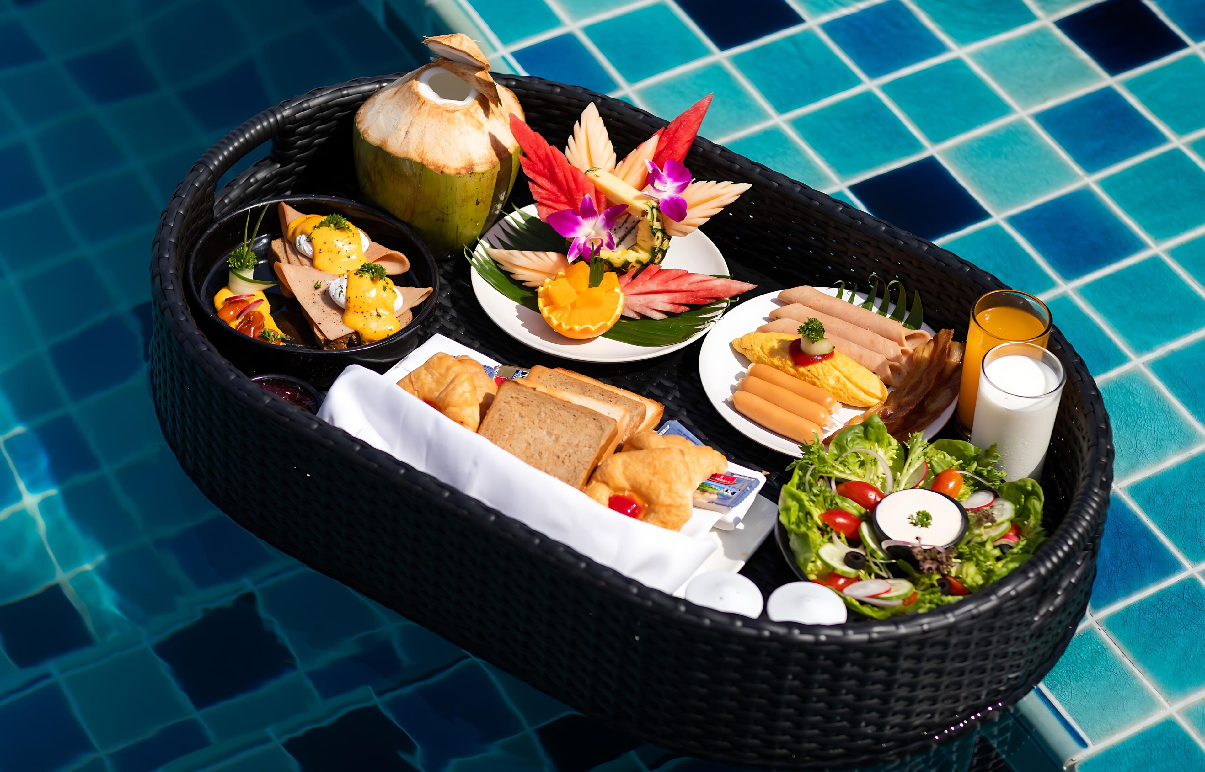 A food basket with various dishes and drinks by a pool.