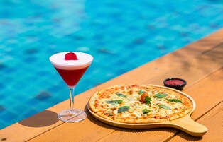 Una mesa de madera con pizza y una copa de martini junto a una piscina.