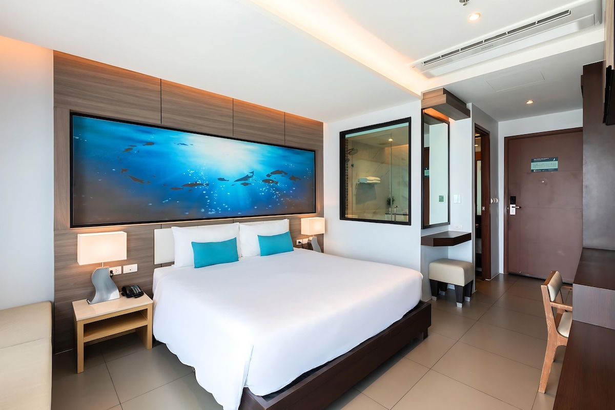 A hotel room with a large bed, a nightstand, a desk, a chair, a large mirror, and a wall-mounted TV.