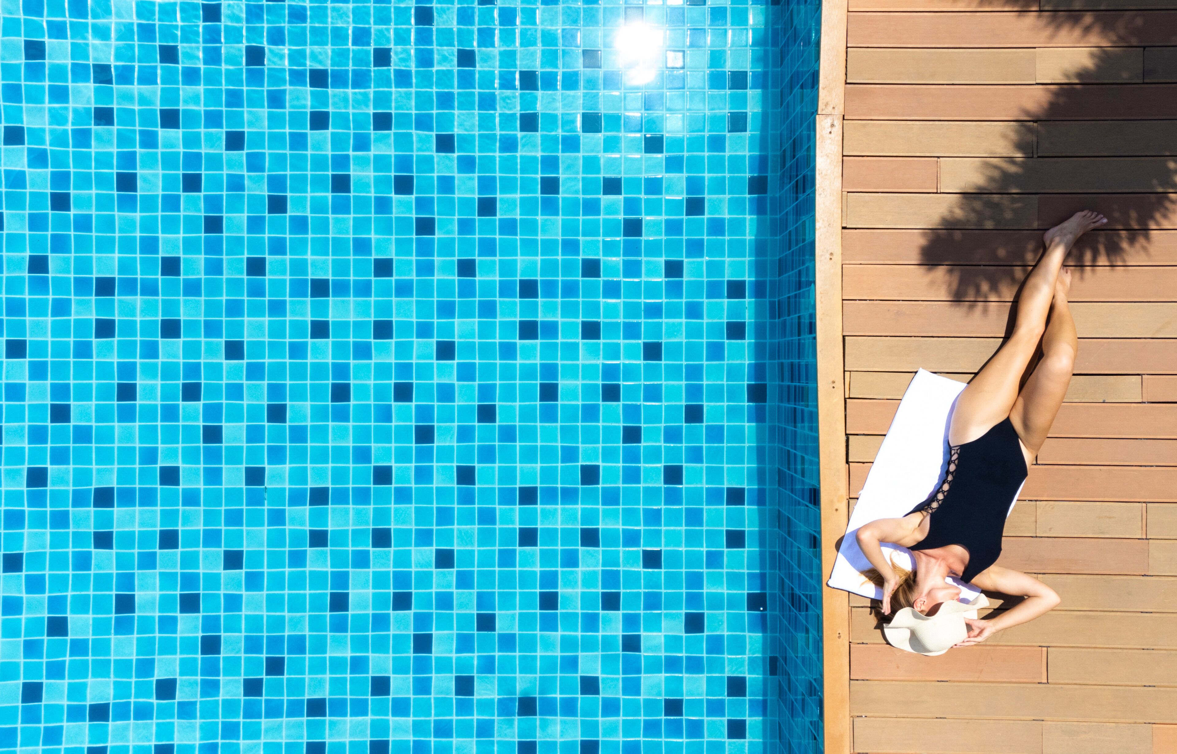 A person lying on a sun lounger by a swimming pool with a tiled floor.