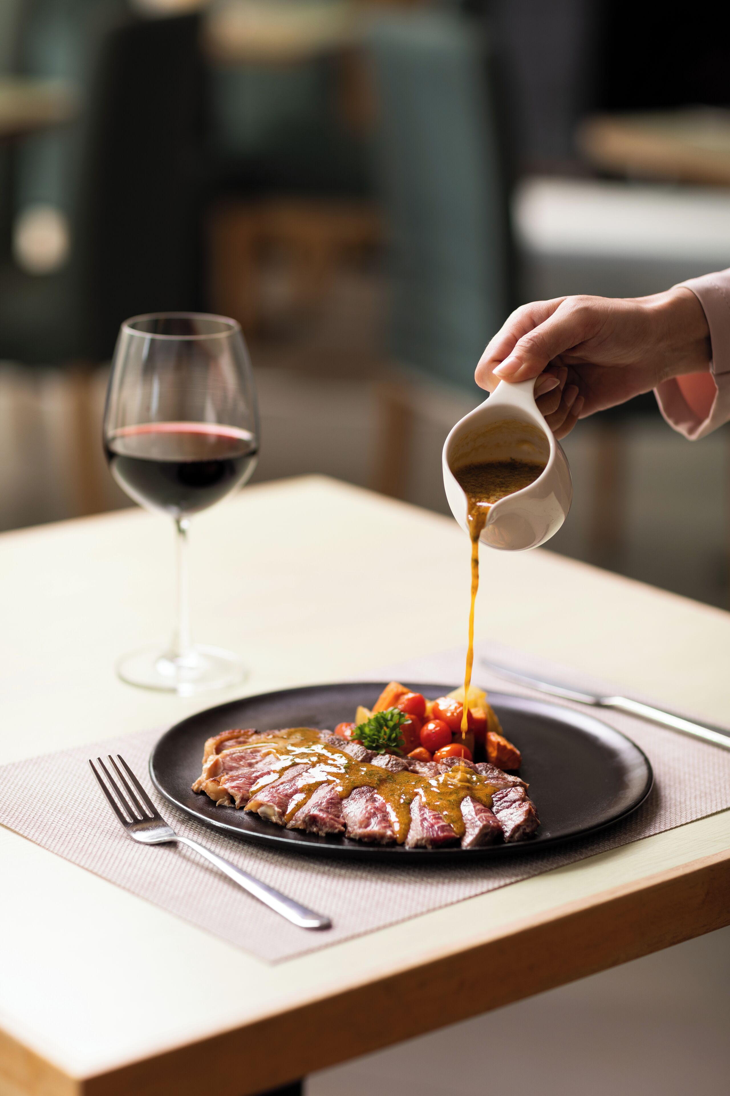 A steak with sauce on a plate, a glass of red wine, and a fork on a table.