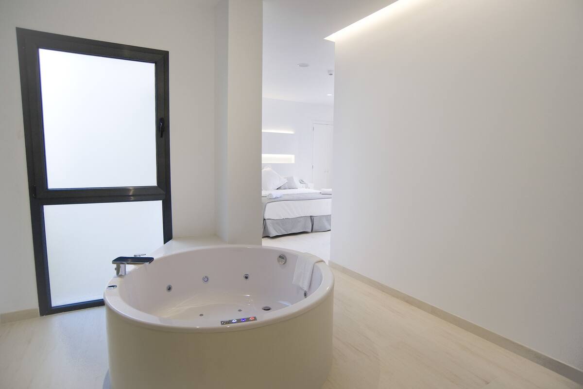 Une salle de bain moderne avec une grande baignoire, une fenêtre et une chambre à coucher visible au fond.