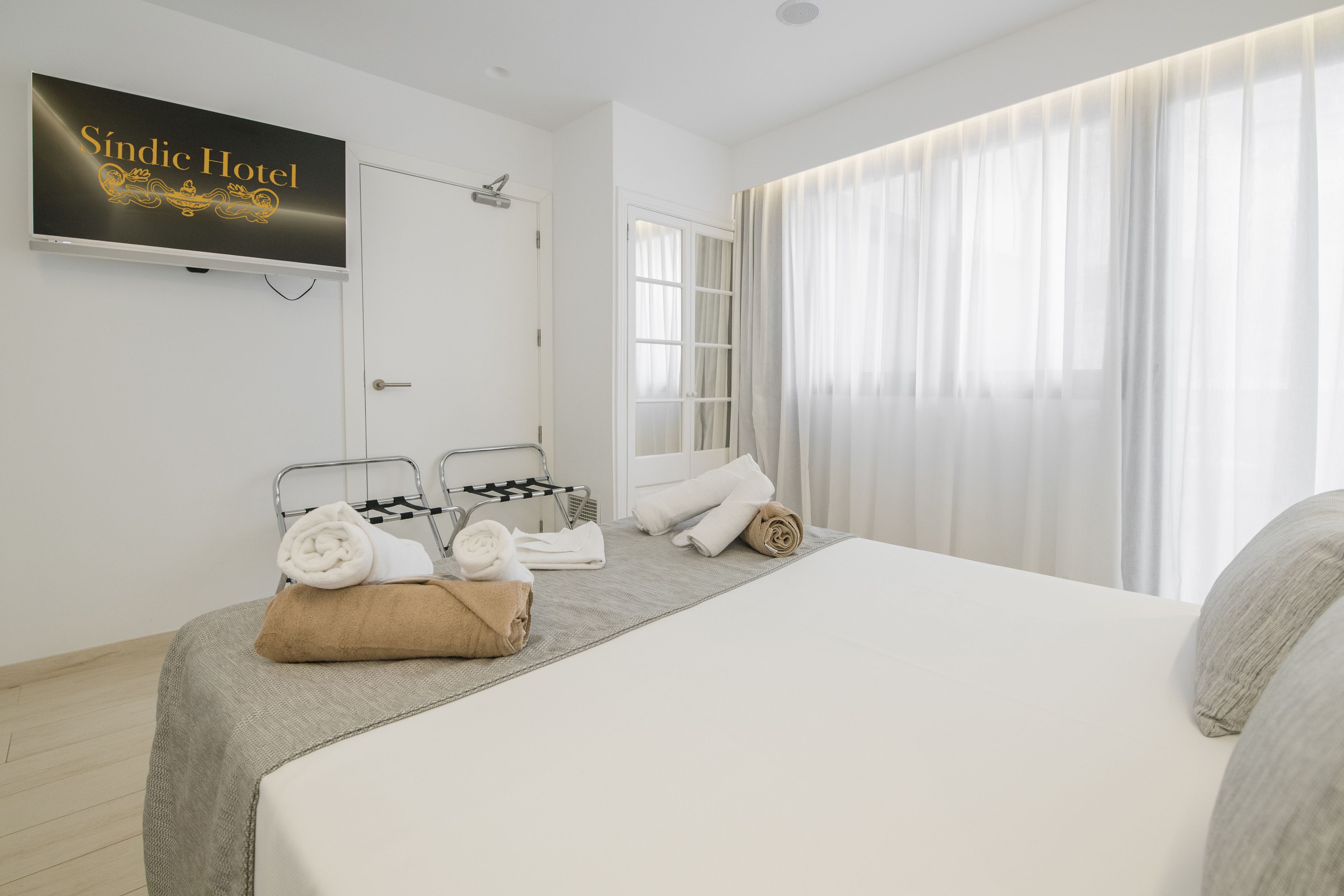 Habitación de hotel con cama, televisor mostrando 'Sídic Hotel', ventana con cortinas y toallas sobre la cama.