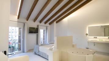 A modern bedroom with a large bed, a sitting area, a balcony, and a bathroom with a bathtub and a large mirror.