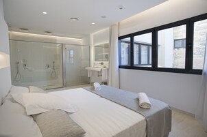 Una habitación de hotel moderna con una cama grande, una ducha con cerramiento de vidrio, un baño con espejo y una ventana que ofrece vistas al exterior.