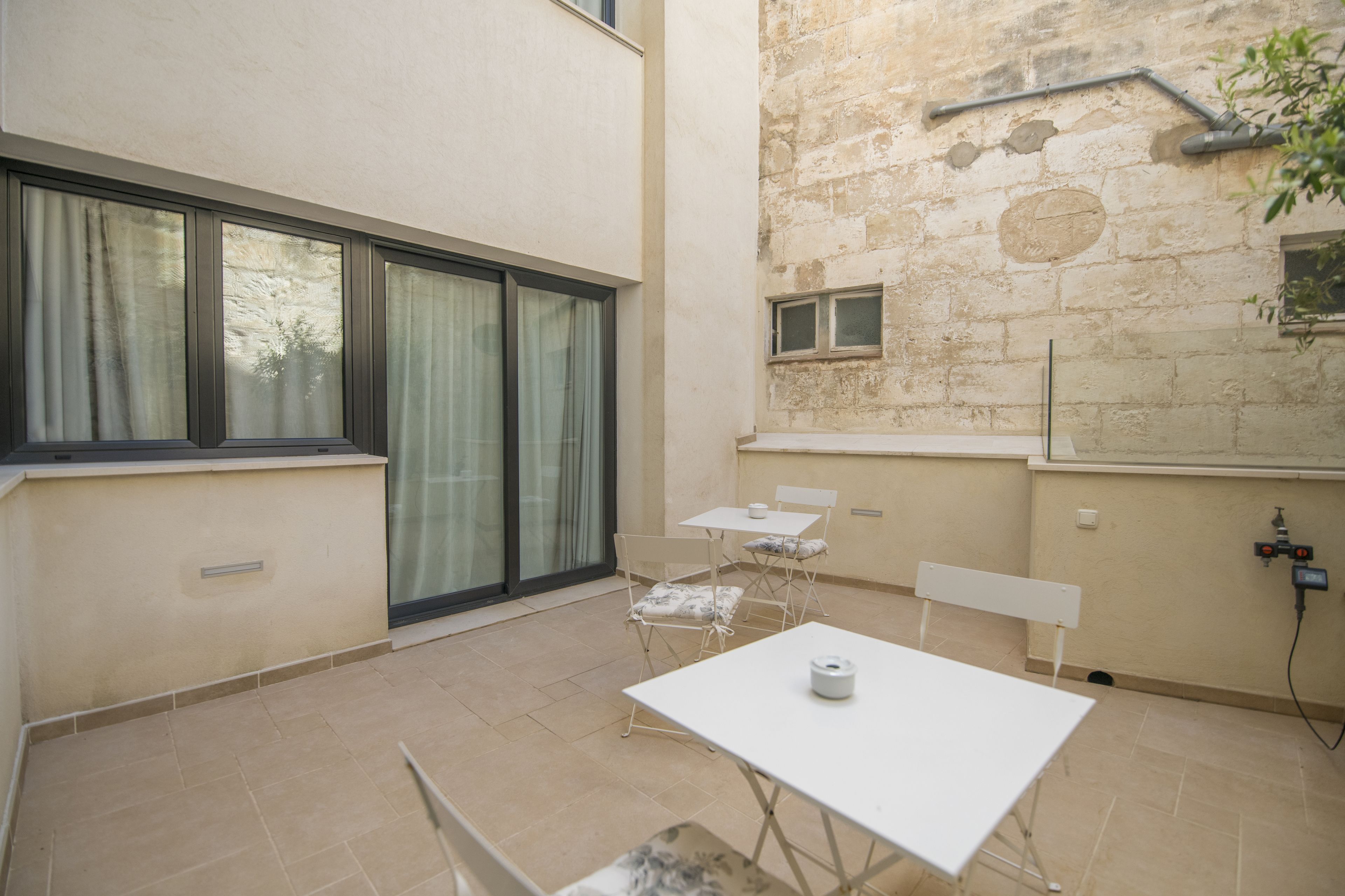 Ein kleiner, offener Wohnbereich mit einem weißen Tisch und zwei Stühlen, einem großen Fenster mit Vorhängen und einer Steinwand mit kleinem Fenster.
