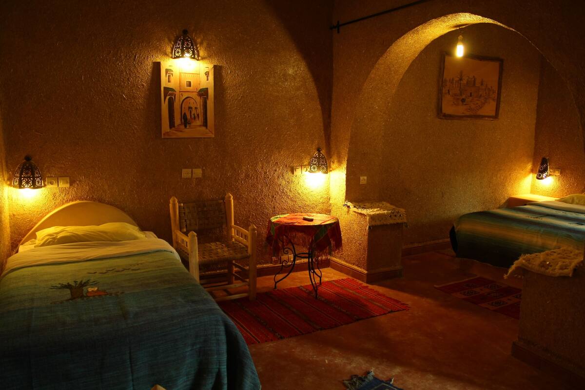 Una habitación tenuemente iluminada con dos camas, una silla, una mesita y luces en la pared.