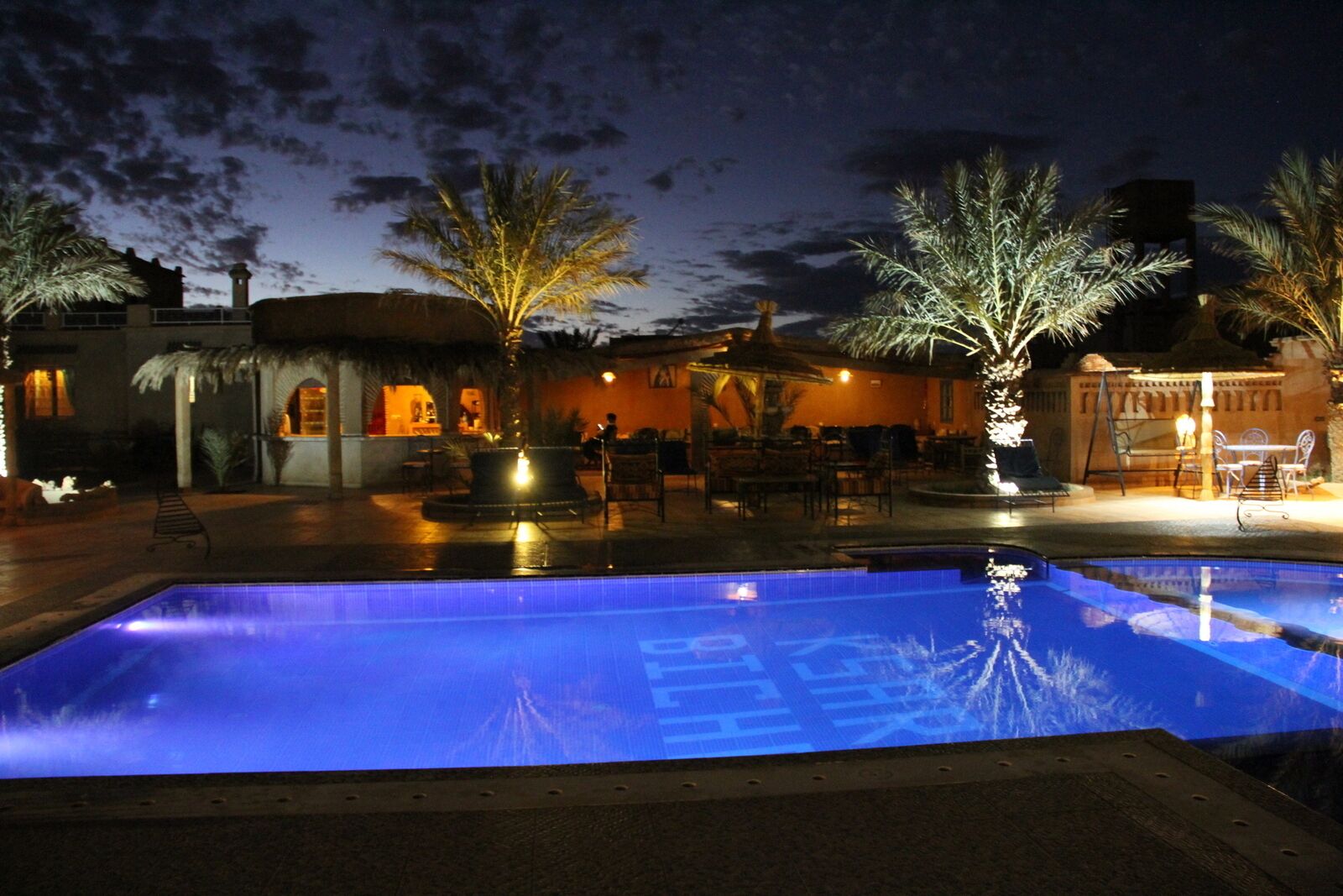 Zona de piscina nocturna con una piscina azul cristalina, palmeras y mobiliario exterior.