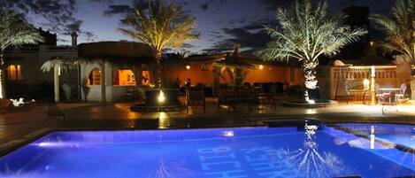Zona de piscina nocturna con una piscina azul cristalina, palmeras y mobiliario exterior.