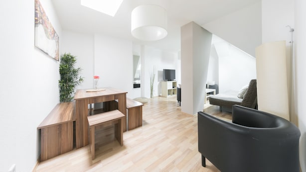 Ein modernes Wohnzimmer mit einem Holztisch, einem schwarzen Sessel und einer Couch. An der Wand befinden sich ein Fernseher und eine Pflanze.