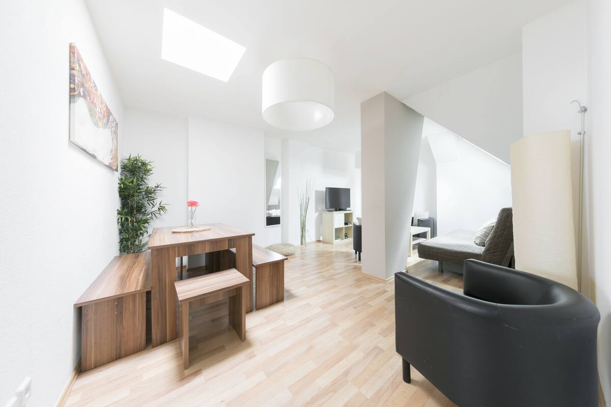 Una sala de estar moderna con un escritorio de madera, un sillón negro y un sofá. Hay un televisor y una planta en la pared.