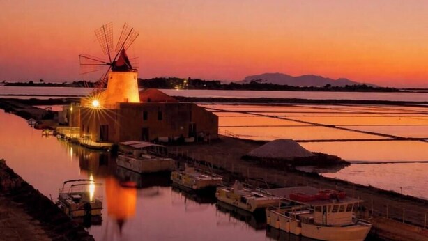 A windmill, boats, and a building by a calm water body at sunset. - Baglio il Giovinetto (Marsala)