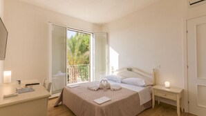 A bedroom with a bed, bedside table, and a desk with a chair. There is a balcony with a view of greenery. - Baglio il Giovinetto (Marsala)
