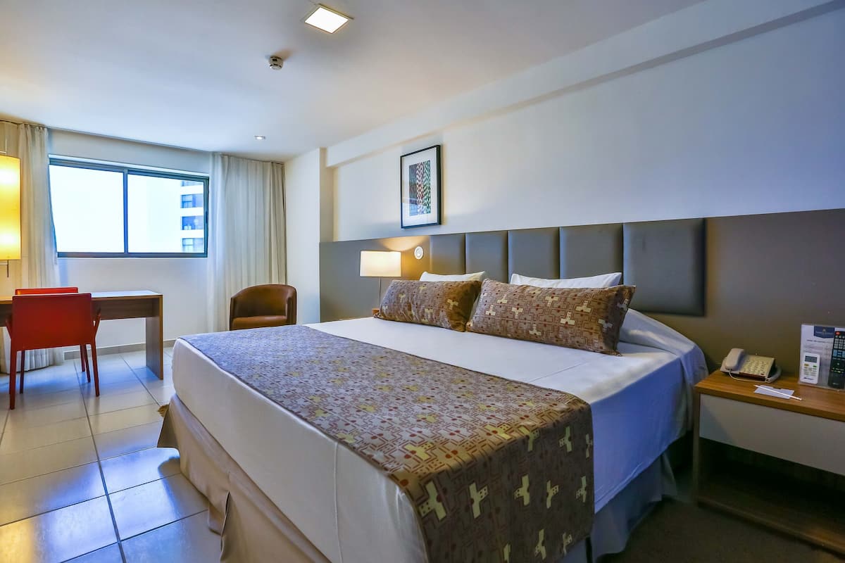 Une chambre d’hôtel avec un grand lit, un bureau, une chaise, une table de chevet avec un téléphone et une fenêtre avec des rideaux.