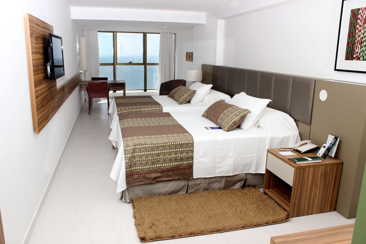 Une chambre d’hôtel avec deux lits, une télévision, un bureau avec une chaise, une fenêtre donnant sur un paysage agréable et un petit tapis.