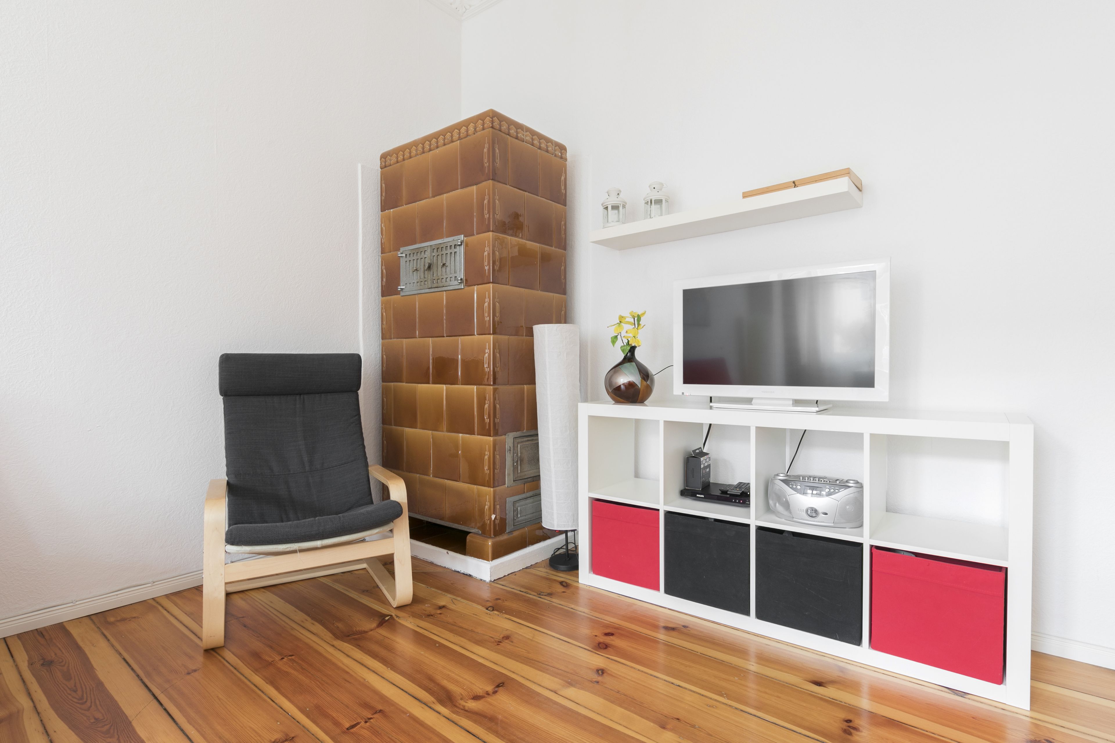 Un salón con piso de madera, una silla negra, un mueble blanco de entretenimiento con compartimentos de almacenamiento rojo y negro, un televisor de pantalla plana y un jarrón decorativo con flores.